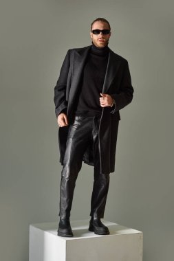 Modayı takip eden güneş gözlüklü ve ceketli modacı erkek model gri kübün üstünde duruyor.