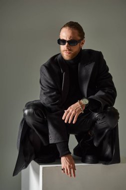 fashionable male model in trendy sunglasses and coat sitting on top of on white cube on grey clipart