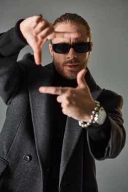 portrait of handsome male model in trendy sunglasses and black attire showing frame gesture on grey clipart