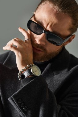 pensive and bearded man in trendy dark sunglasses and black attire posing on grey background clipart