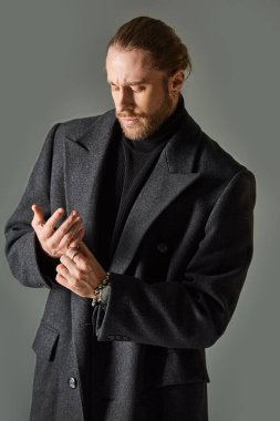 good looking and bearded man in trendy black attire and woolen coat posing on grey background clipart