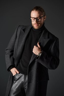 handsome man in eyeglasses and classy woolen coat posing with hand in pocket on grey background clipart