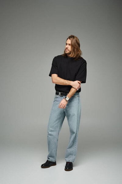 full length of bearded handsome man in black t-shirt and jeans standing on grey background