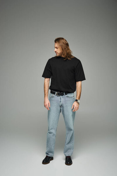 full length of bearded handsome man in black t-shirt and jeans standing on grey background