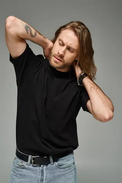 bearded and handsome man with long hair posing in jeans and t-shirt with hands behind neck on grey