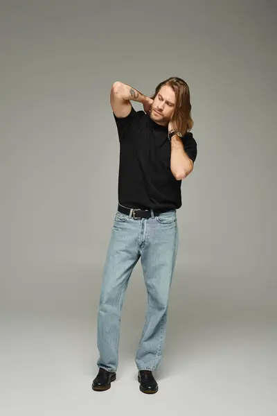 bearded and handsome man with long hair posing in jeans and t-shirt with hands behind neck on grey