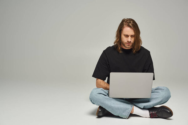 handsome man with long hair and beard sitting with crossed legs on floor and using laptop on grey