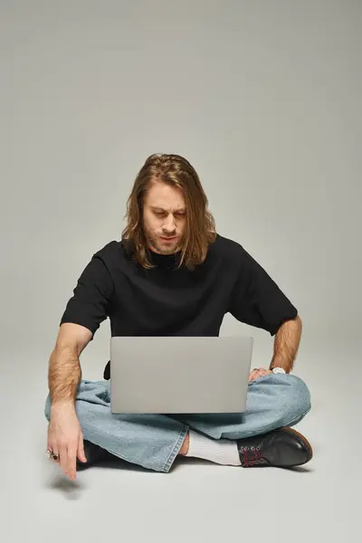 handsome man with long hair and beard sitting with crossed legs on floor and using laptop on grey