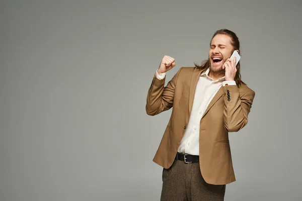 stock image handsome and excited bearded businessman in formal attire talking on smartphone on grey backdrop