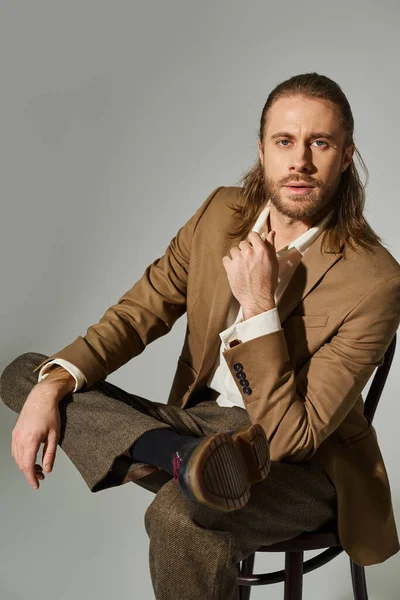 stock image handsome bearded businessman in beige formal attire sitting on wooden chair on grey background