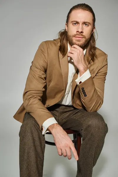 stock image handsome bearded businessman in beige formal attire sitting on wooden chair on grey background