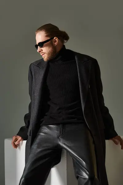 stock image portrait of handsome man in trendy sunglasses and woolen coat sitting on white cube on grey backdrop