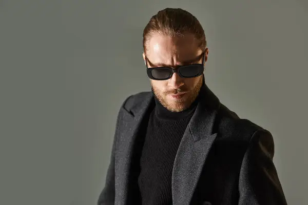 stock image portrait of handsome bearded man in trendy sunglasses and black attire posing on grey backdrop