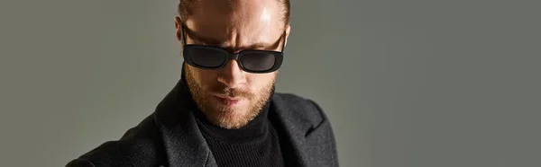 stock image portrait of bearded man in trendy sunglasses and black attire posing on grey backdrop, banner