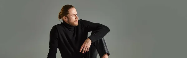 stock image banner of bearded man in turtleneck sweater and black leather pants sitting on grey background