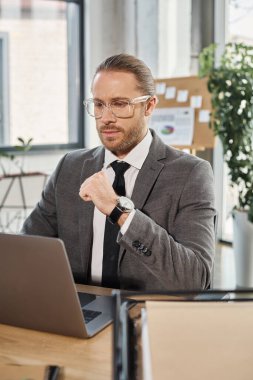 Gri takım elbiseli ve gözlüklü düşünceli bir işadamı modern ofis alanında dizüstü bilgisayarla çalışıyor.