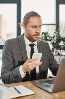 Gri takım elbiseli sakallı iş adamı elinde akıllı telefon ve ofisteki bilgisayarına bakıyor.