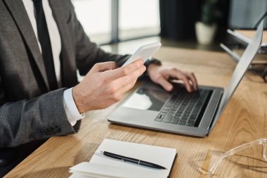 Bilgisayarın yanında akıllı telefonu ve çalışma masasının üstünde not defteri olan iş adamının kırpılmış görüntüsü