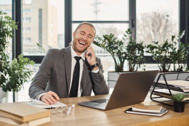 heyecanlı başarılı iş adamı akıllı telefonda konuşuyor ve modern ofiste dizüstü bilgisayarın yanında gülüyor