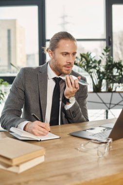 İş adamı akıllı telefondan sesli mesaj gönderiyor ve ofisteki dizüstü bilgisayarın yanında not defteri yazıyor.