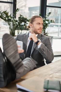 relaxed successful businessman in grey suit sitting with legs on desk and looking away in office clipart