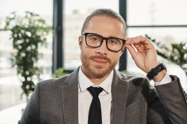 Gri takımlı başarılı girişimci gözlükleri ayarlıyor ve ofisindeki kameraya bakıyor.