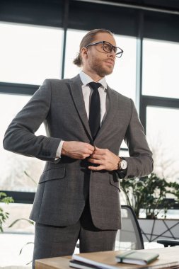 confident businessman in eyeglasses buttoning stylish blazer and looking away in modern office clipart