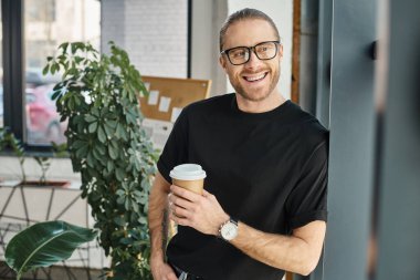 cheerful businessman in black t-shirt and eyeglasses holding coffee to go and looking away in office clipart