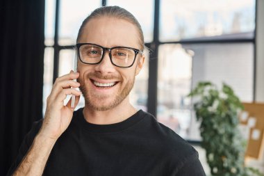 Gözlüklü mutlu iş adamının portresi akıllı telefonla konuşuyor ve ofisteki kameraya bakıyor.