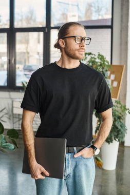 corporate manager in black t-shirt and eyeglasses standing with laptop and hand in pocket in office clipart