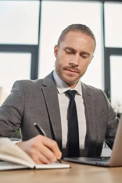 stock image serious and ambitious businessman in grey sitting near laptop and writing in notebook, productivity