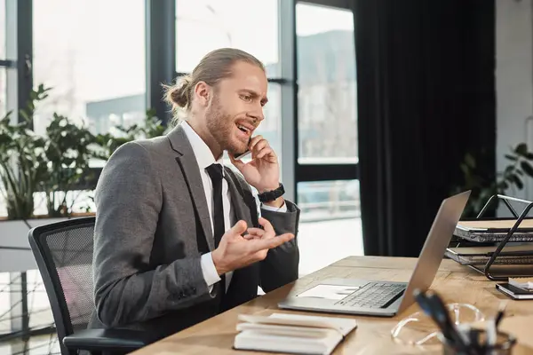 nervous businessman pointing at laptop while talking on mobile phone at workplace in office
