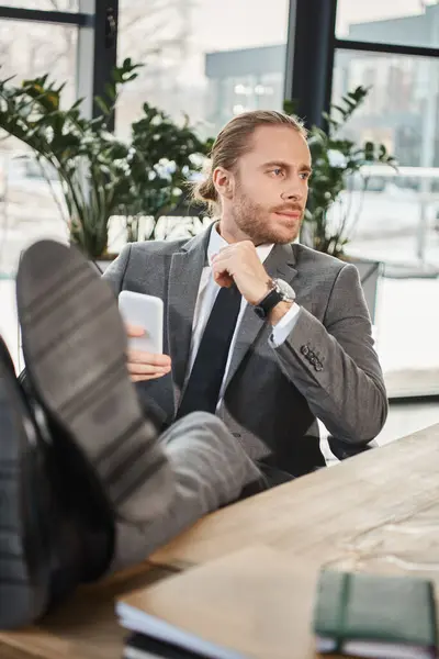 relaxed successful businessman in grey suit sitting with legs on desk and looking away in office