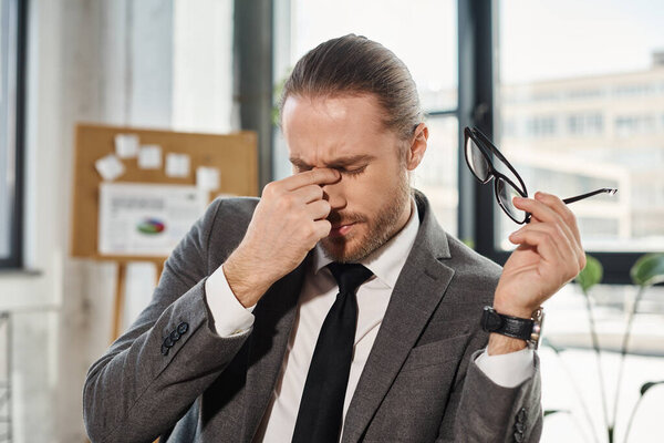 tired businessman in formal wear holding eyeglasses and rubbing eyed while sitting in office