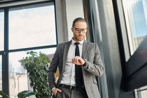 stylish businessman in grey suit and eyeglasses standing in modern office and looking at wristwatch