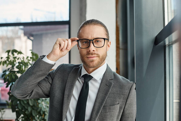 portrait of entrepreneur in formal wear adjusting eyeglasses and looking at camera in office