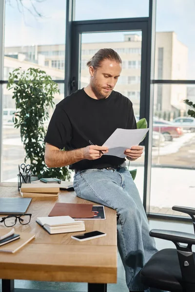 stock image manager in casual attire sitting on work desk near notebooks and looking at documents, paperwork