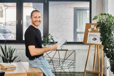 cheerful manager holding documents and looking at camera near work desk in contemporary office clipart