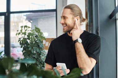 Siyah tişörtlü neşeli iş adamı akıllı telefon tutuyor ve ofiste kulaklıkla müzik dinliyor.