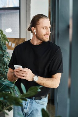 Siyah tişörtlü mutlu iş adamı akıllı telefon tutuyor ve ofiste kulaklıkla müzik dinliyor.
