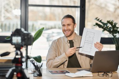 cheerful businessman showing blueprint in front of digital camera during video blog in modern office clipart