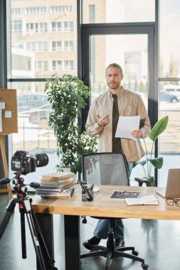 serious businessman holding document and recording video blog in front of digital camera and laptop clipart