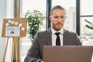 confident manager in eyeglasses and grey suit working on laptop in modern office environment clipart