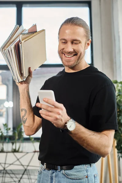 joyous businessman in casual attire holding pile of notebooks and messaging on smartphone in office