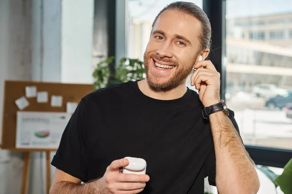 stock image cheerful businessman in casual attire adjusting wireless earphone and smiling at camera in office