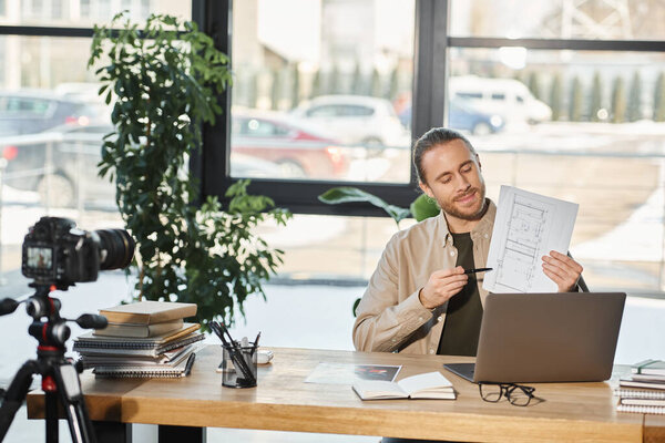 бизнесмен в повседневной одежде показывает чертеж во время видео блога на ноутбуке рядом с цифровой камерой
