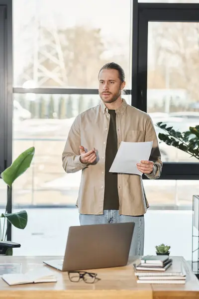 stock image serious businessman holding document and talking during video conference on laptop in office