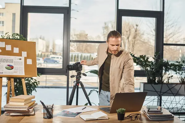 stock image manager in casual attire adjusting laptop and digital camera near work desk in office, video blogger