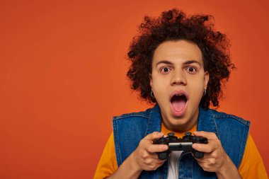 attractive african american man in casual attire emotionally playing video games on orange backdrop clipart