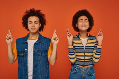 young attractive african american siblings posing together and making wish on orange backdrop clipart